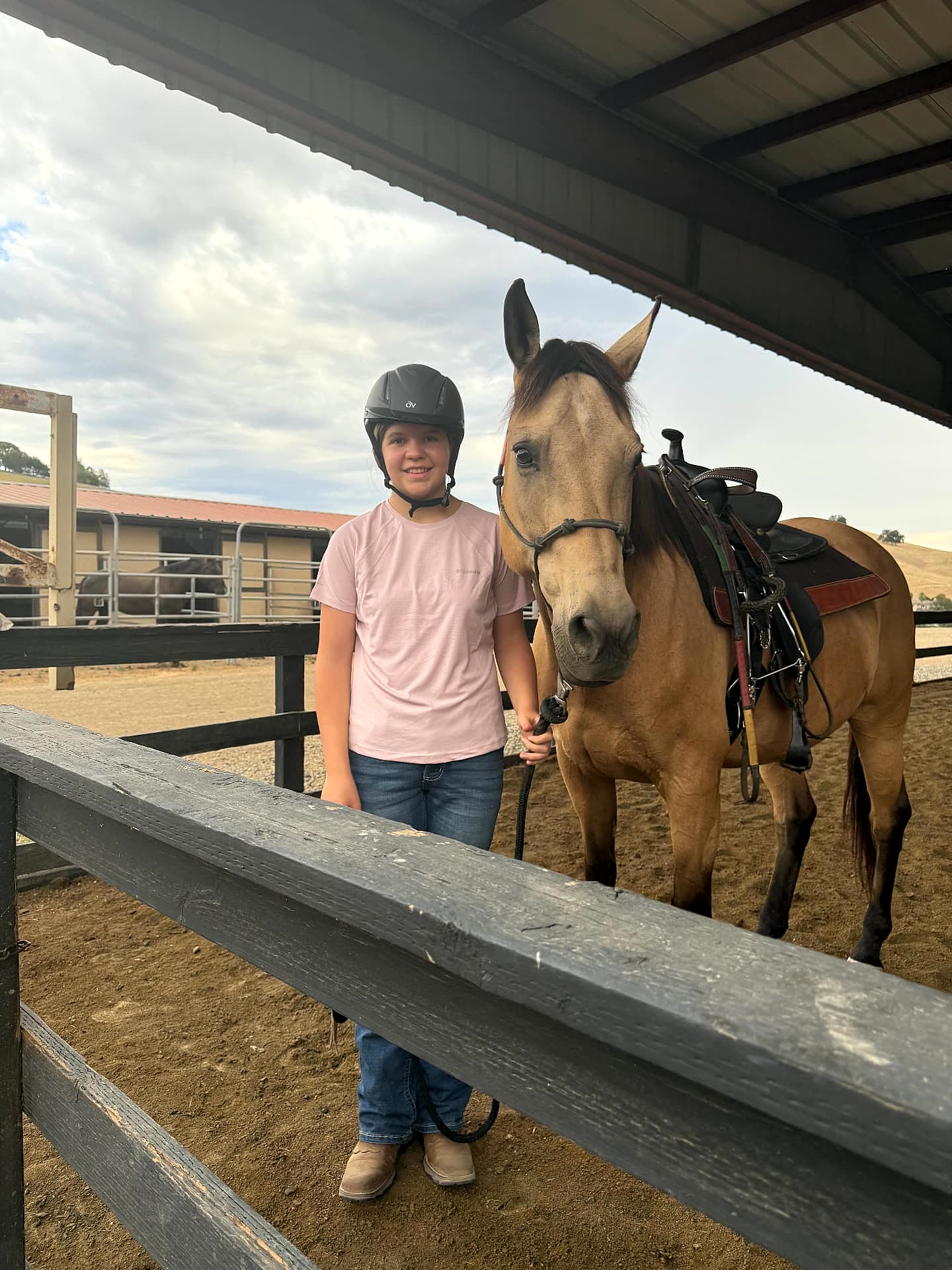 Young rider in a helmet standing with her buckskin horse in the covered arena
